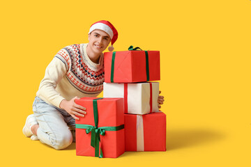 Happy young man in Christmas sweater and Santa hat with gift boxes sitting on yellow background