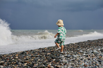 A toddler in a cute outfit and a sunhat walks along a rocky beach near the ocean as waves crash...