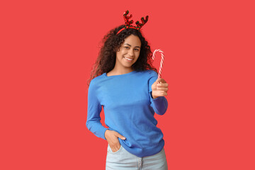 Beautiful young African-American woman in reindeer horns with candy cane on red background. Christmas celebration