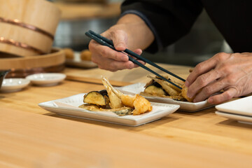 Obraz premium Japanese chef carefully plating tempura vegetables and seafood using chopsticks, creative presenting the dish on a rectangular ceramic plate in a kitchen setting