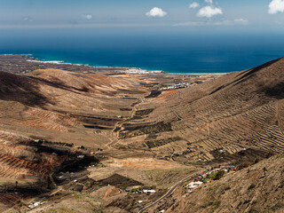 The volcanic landscape of Lanzarote presents a rugged terrain with reddish-brown hills, sparse vegetation, and winding roads. White houses contrast against the barren earth, with the ocean visible in 