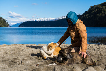 A man in outdoor gear sits on a lakeside beach petting two dogs on a sunny day. The background showcases a pristine lake surrounded by forested mountains with snow-capped peaks.