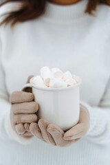 Woman holding in hands hot drink with marshmallow in white ceramic cup in snowy winter forest