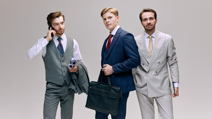 Young businessmen standing confidently, with one on phone and others holding briefcases and documents. Moment of multitasking against white background. Concept of fashion, style, beauty, aesthetic. Ad