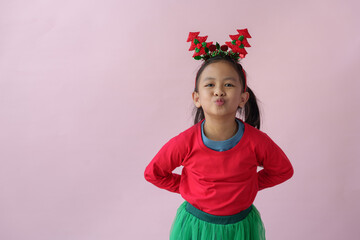 Adorable little Asian girl with Christmas tree headband, antlers showing various poses, standing, smiling, laughing, having fun in various poses, pink background. Holiday concept annual festival.
