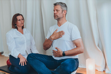 A man lying on a mat with a blindfold, practicing breathing exercises while a woman acts as a...
