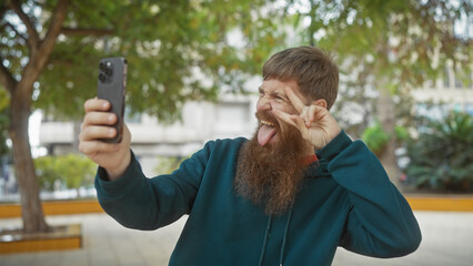 Bearded man taking a playful selfie outdoors with greenery in the background.