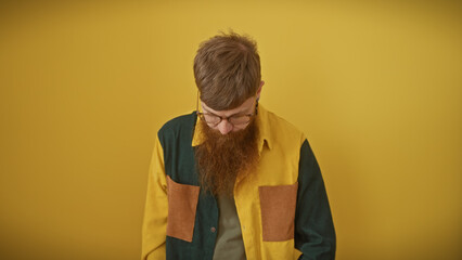 A thoughtful young man with a redhead and beard posing over a yellow background wearing glasses and...