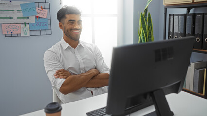 Naklejka premium Young man working at an office desk, smiling at his computer with arms crossed, surrounded by organizational charts and a cup of coffee