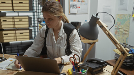 Focused woman detective working indoors at a cluttered police office, analyzing data on her laptop...