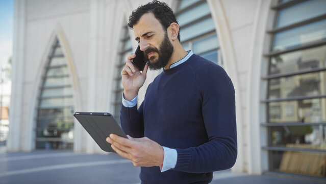 Handsome middle-aged man with beard using smartphone and tablet on city street outdoor.