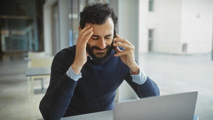 Stressed man on phone in office with laptop, expressing frustration and discomfort accompanied by...