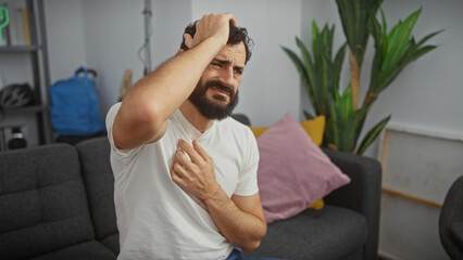 Middle-aged bearded man in pain holding head in a modern living room setting, expressing discomfort.