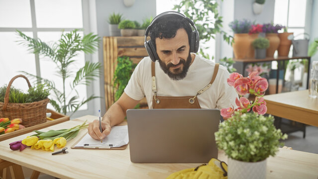 Bearded man in headphones working on laptop at a flower shop with plants and clipboard on wooden table.