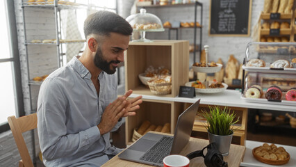 Young hispanic man working on laptop in a cozy bakery interior filled with assorted pastries and desserts