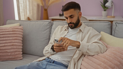 Young hispanic man with beard using smartphone in modern living room setting, showcasing casual indoor lifestyle with comfortable home decor