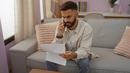 Young man with a beard sitting on a couch in a living room, looking concerned while talking on the phone and holding papers.