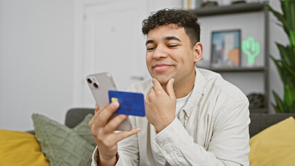 Handsome man holding credit card and smartphone contemplating online shopping at home.