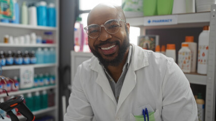 Handsome young african american man with a beard smiling in a pharmacy setting, showcasing assorted products on shelves indoors