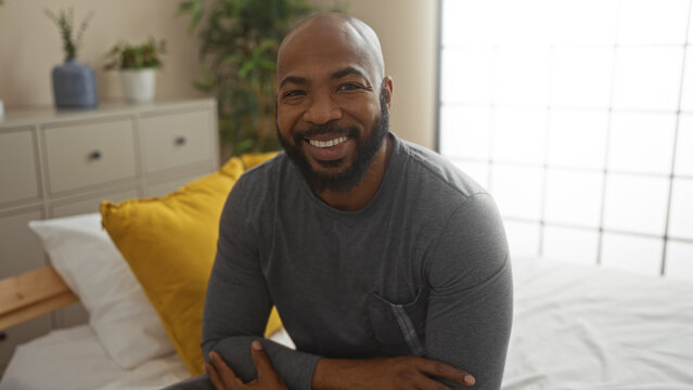 Young man with beard sitting on bed in cozy bedroom with plants and pillows, smiling confidently indoors at home - Powered by Adobe