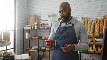 Young man inside a bakery shop checking his phone while holding a credit card under bright natural...