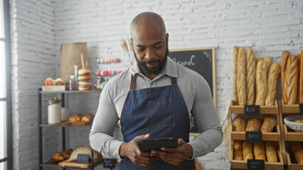 Young man in bakery wearing apron and using tablet in a bright indoor setting with shelves full of...