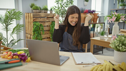 Young hispanic woman celebrating success in a flower shop with laptop