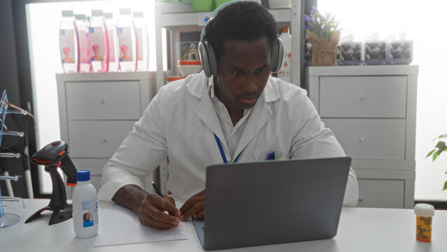 Handsome young man taking notes in a pharmacy store, sitting indoors using a laptop, wearing headphones and a white lab coat. - Powered by Adobe