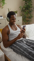 Handsome man relaxing on bed in bedroom at home wearing headphones and using smartphone, surrounded by indoor plants