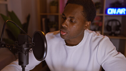 A focused african american man recording in a cozy, dimly-lit room with an 'on air' neon sign.