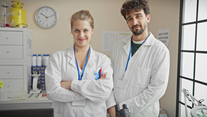 A woman and a man in lab coats stand confidently indoors in a well-equipped laboratory environment, implying a professional clinical setting.