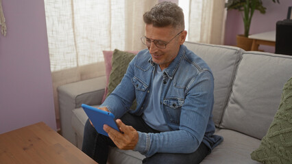 Middle-aged man reading tablet on couch in living room of cozy home interior