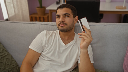 Obraz premium Young hispanic man holding a credit card while relaxing on a sofa in the living room of a cozy apartment, showcasing a calm and thoughtful demeanor.