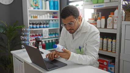 Young man in white coat examining product in pharmacy while using laptop in a bright and well-organized store with various medications and bottles on shelves