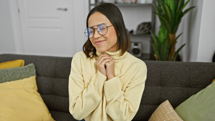 Smiling hispanic woman with glasses wearing a yellow sweater seated on a grey couch indoors.