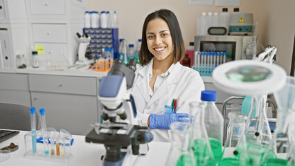 A smiling young hispanic woman in a lab coat working with a microscope in a well-equipped laboratory setting.