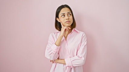 A thoughtful young hispanic woman in a pink shirt poses against a pink background, embodying adult...