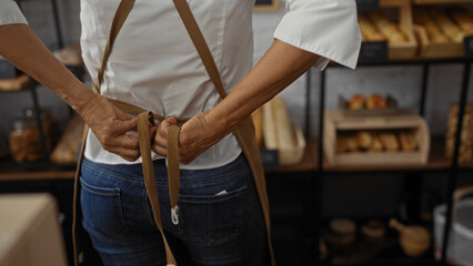 Woman tying apron in a bakery, showcasing fresh bread and pastries on shelves in an indoor shop setting.
