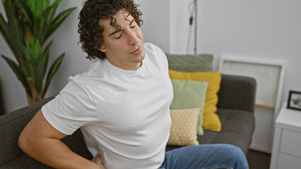 Fototapeta premium A young hispanic man grimaces with back pain while sitting indoors in a living room, adding a human element to the home environment.