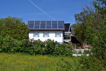 Solar or photovoltaic panels installed on the roof of a white family house observed during sunny day in a Swiss village. There are trees and bushes around the house.