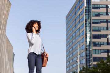 cheerful ethnic female entrepreneur standing in downtown and having conversation on mobile phone while looking away