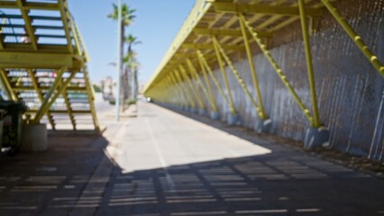 Blurred view of outdoor urban bridge emphasizing yellow metal structure with defocused pedestrian crossing and palm trees under clear sky