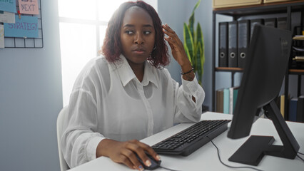 Woman working at a computer in a modern office, appearing thoughtful and focused while surrounded by organizational tools and files.