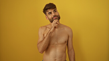 Fototapeta premium Shirtless young hispanic man with a beard standing thoughtfully against a yellow background