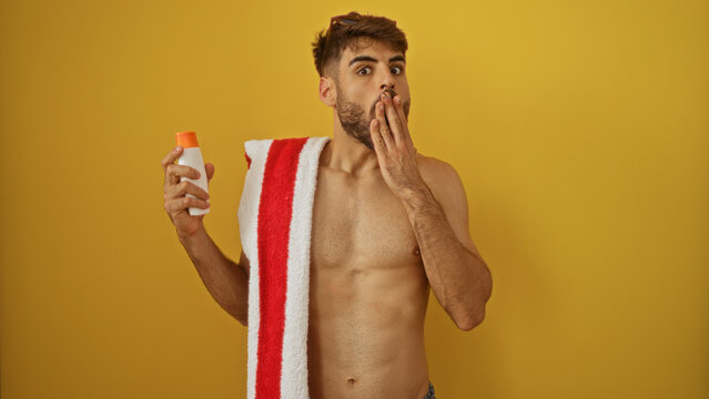 Handsome young hispanic man holding sunscreen with a towel over shoulder standing against yellow background looking surprised