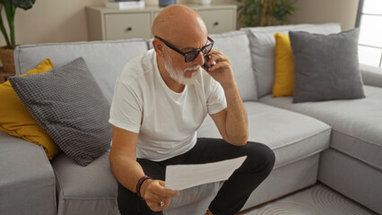 Fototapeta premium Mature man with beard and glasses sitting on a sofa in his living room talking on the phone and holding a document.
