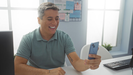 Young man smiling in an office while holding a smartphone, sitting at a white desk with a computer...
