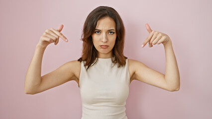 Fototapeta premium A confident young hispanic woman gesturing towards herself against a solid pink background, exuding self-assurance.