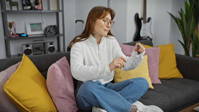 A casual young woman sits on a couch indoors, looking at her smartwatch in a modernly furnished living room.