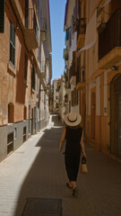 Woman walking through a narrow street in palma, mallorca, spain, wearing a hat and holding a wicker bag, under a clear blue sky in an outdoor setting surrounded by old buildings © Krakenimages.com